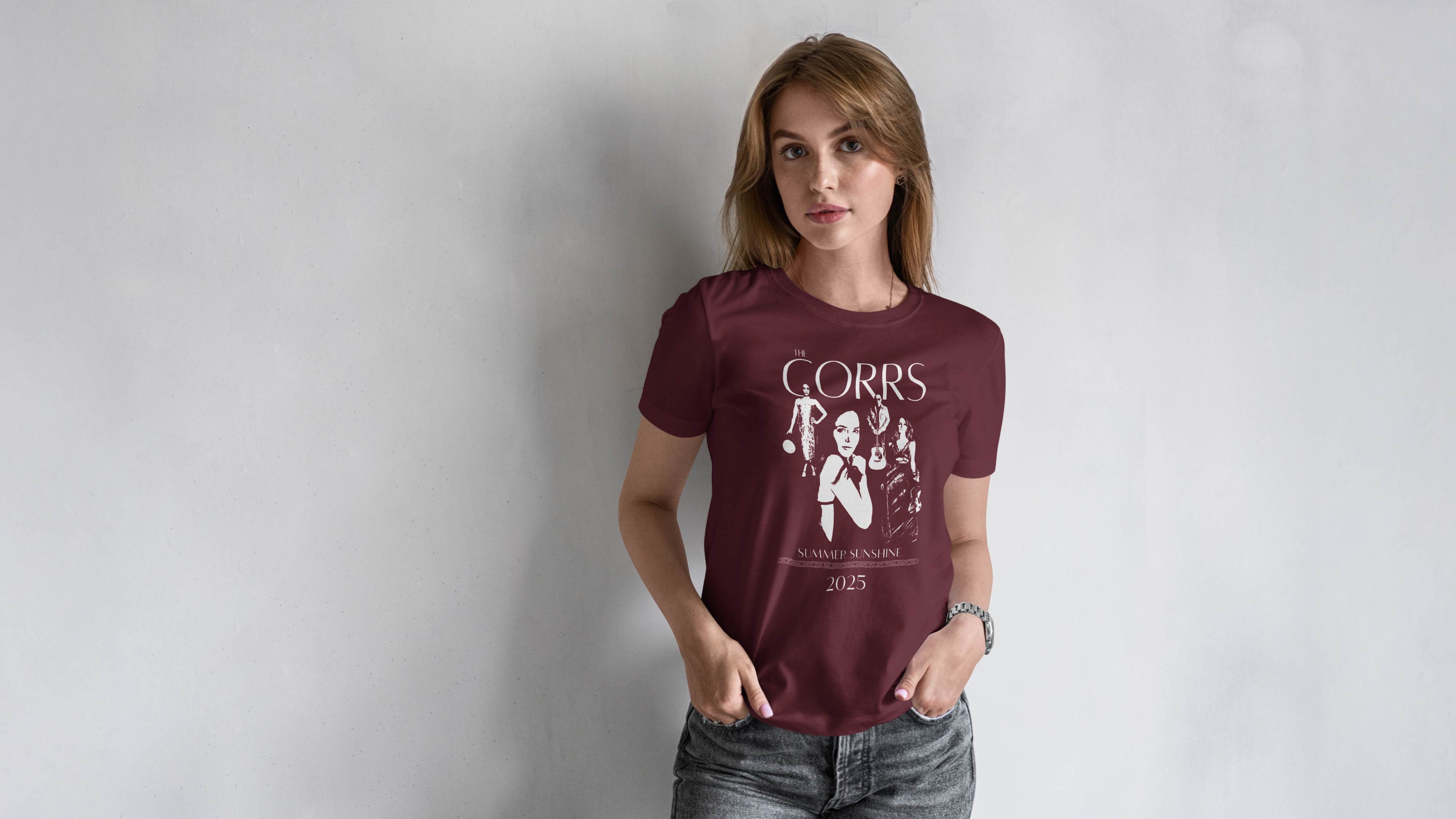Promotional banner for The Corrs' new merchandise release. A young woman stands against a grey wall wearing a maroon T-shirt featuring a white graphic of the band and the text “THE CORRS – SUMMER SUNSHINE 2025.” On the left side, large white text reads “New Releases,” with smaller text below stating, “Be the first to shop The Corrs brand new collection, featuring brand new T-shirts and hoodies!” A green button below says “Explore Now.