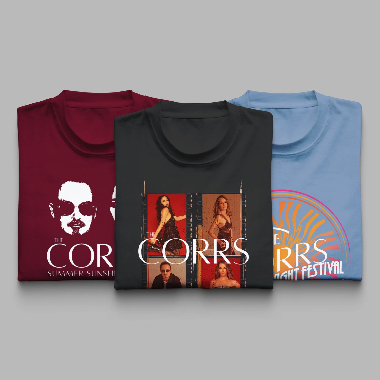 Three folded T-shirts from The Corrs' 2025 tour collection displayed side by side. The left shirt is maroon with a black-and-white graphic of a band member wearing sunglasses and the text “THE CORRS – SUMMER SUNSHINE.” The middle shirt is black, featuring a film strip-style collage of the band members with “THE CORRS” in large white text. The right shirt is light blue with a colourful retro-style sunburst design. All shirts are laid out on a light grey background. 