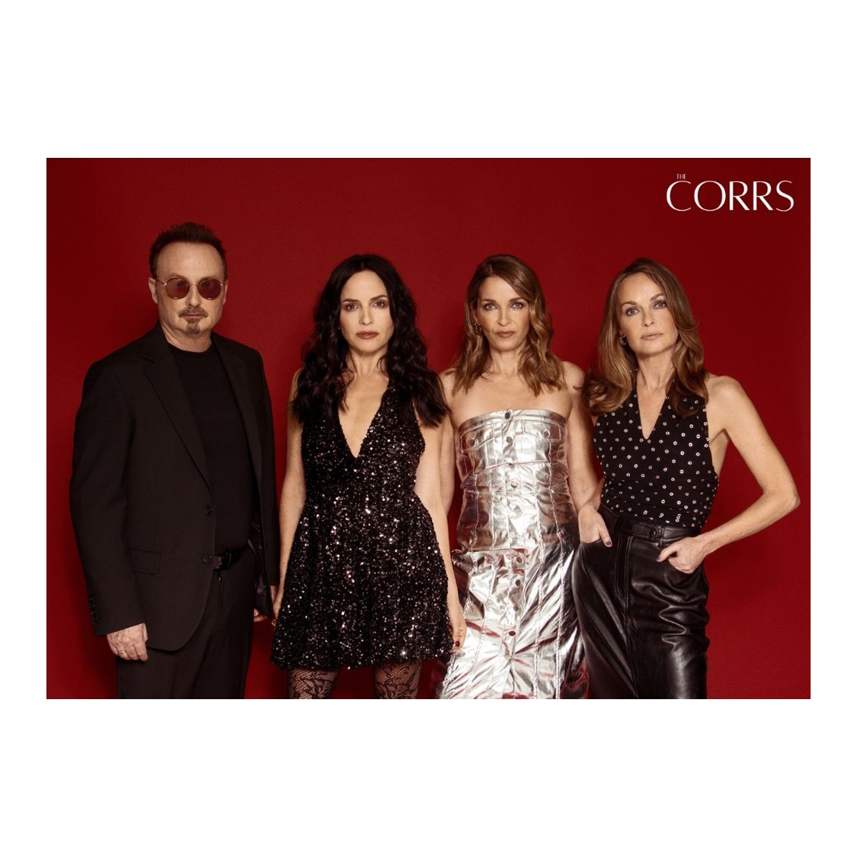 An A3 poster from The Corrs Store by National Merchandise features four people against a red background, with a man in dark sunglasses and black suit, three women in stylish black or metallic outfits, and THE CORRS in white at the top right.