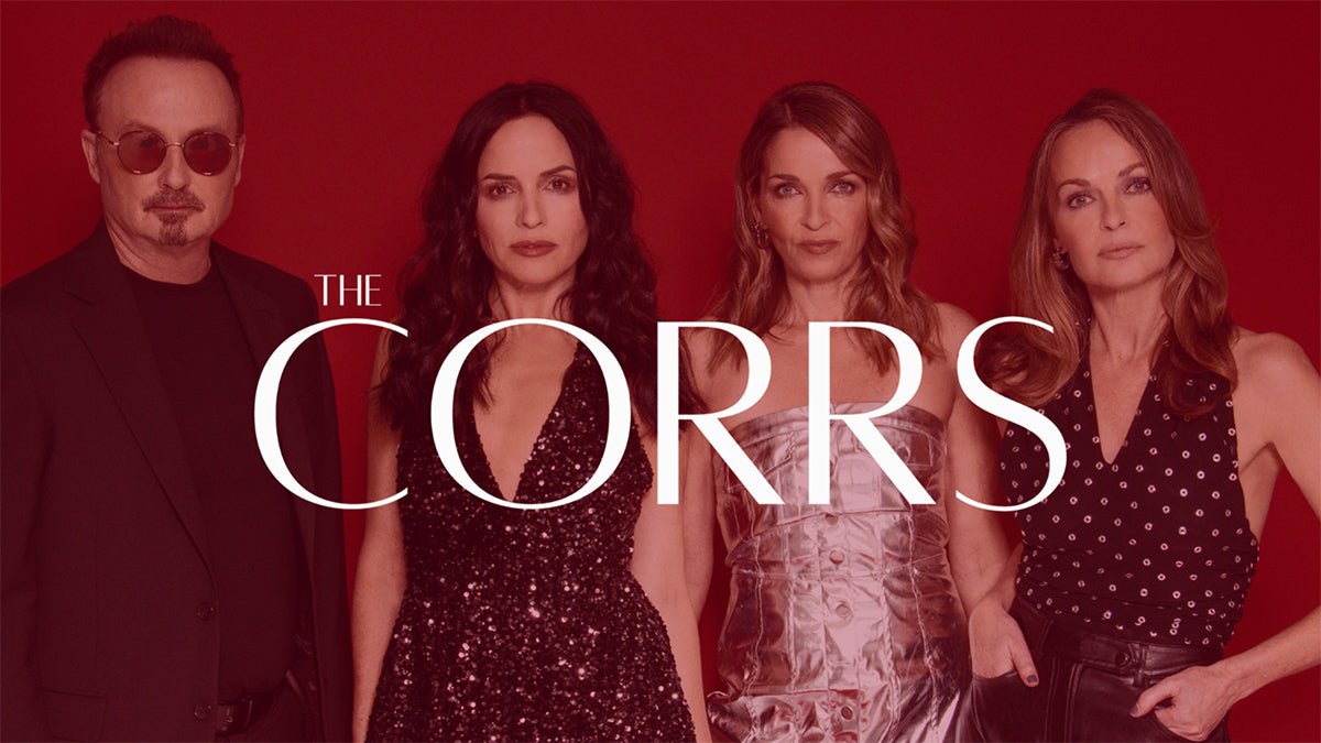 Promotional banner for The Corrs' 2025 tour merchandise. The image shows the four band members standing against a deep red background, dressed in stylish black and metallic outfits. Large white text across the centre reads “THE CORRS,” with a button below stating “Shop 2025 Tour Collection.”